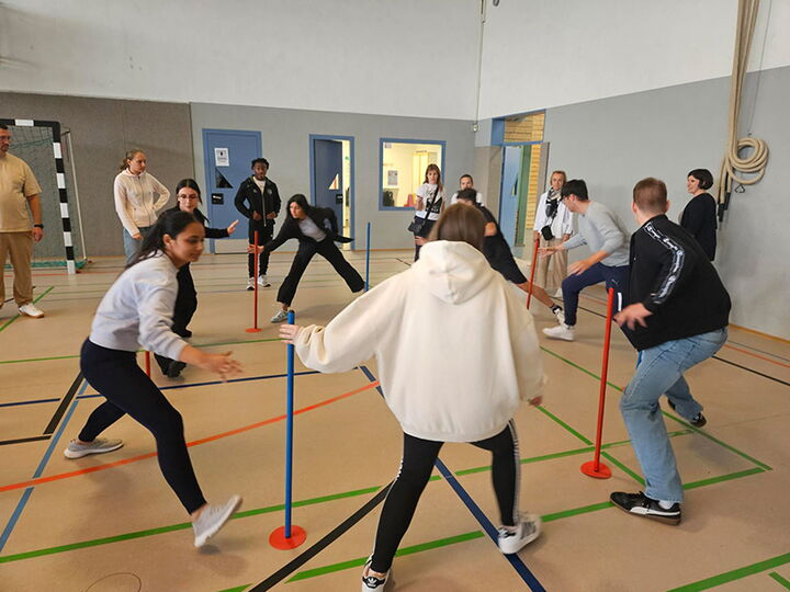 Gruppe junger Menschen spielt ein Bewegungsspiel mit farbigen Stangen in der Sporthalle der Erich-Bracher-Schule.