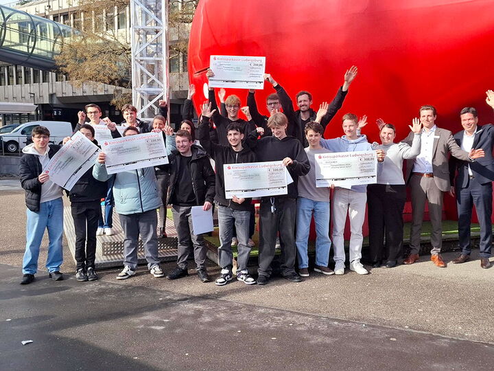 Eine große Gruppe von Schülern, Lehrern und Bankvertretern steht vor dem roten Sparkassen-Sparschwein in Ludwigsburg. Einige halten Zeremonieschecks in die Kamera.