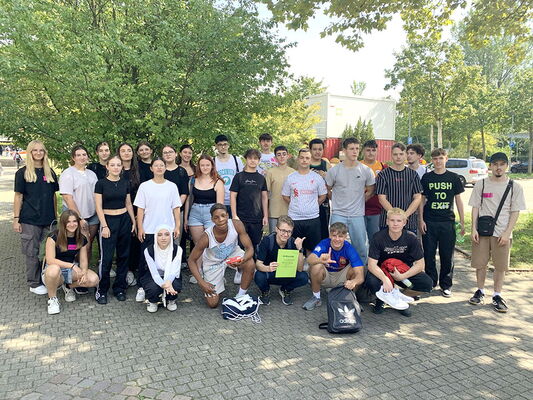 Schulklasse im Freien, aufgenommen im Schatten eines großen Baumes. Einige Schülerinnen und Schüler knien vorn, während andere stehen.