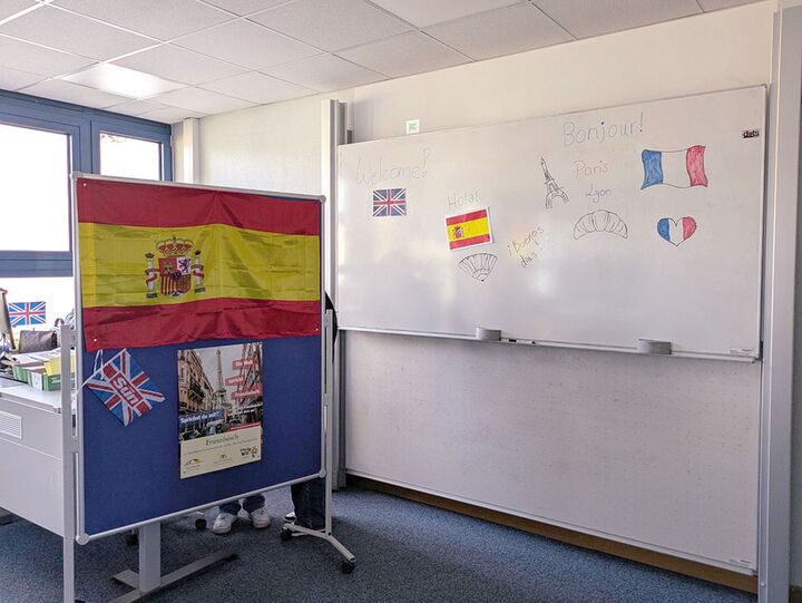 Klassenzimmer mit Pinnwand und spanischer Flagge, daneben eine Tafel mit Begrüßungen auf Englisch, Spanisch und Französisch.