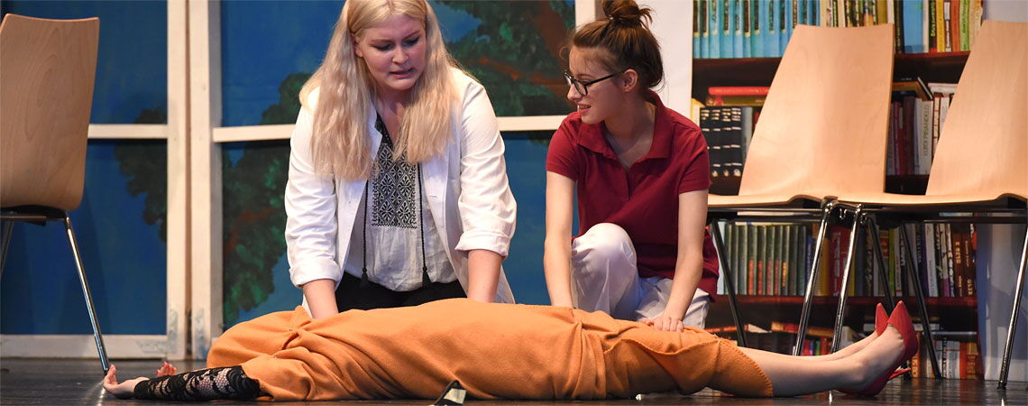 Zwei Schauspielerinnen knien auf der Theaterbühne neben einer Mitspielerin, die in eine orangefarbene Decke gewickelt auf dem Boden liegt.