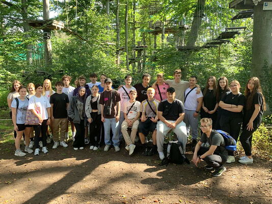 Schüler und Lehrkräfte stehen dicht beisammen im Wald. Im Hintergrund sind Kletterelemente eines Hochseilgartens zu sehen.
