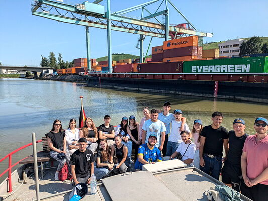 Schülergruppe des Berufskollegs sitzt auf einem Boot im Stuttgarter Hafen. Im Hintergrund sind Container, Kräne und ein Frachtschiff mit der Aufschrift „EVERGREEN“.