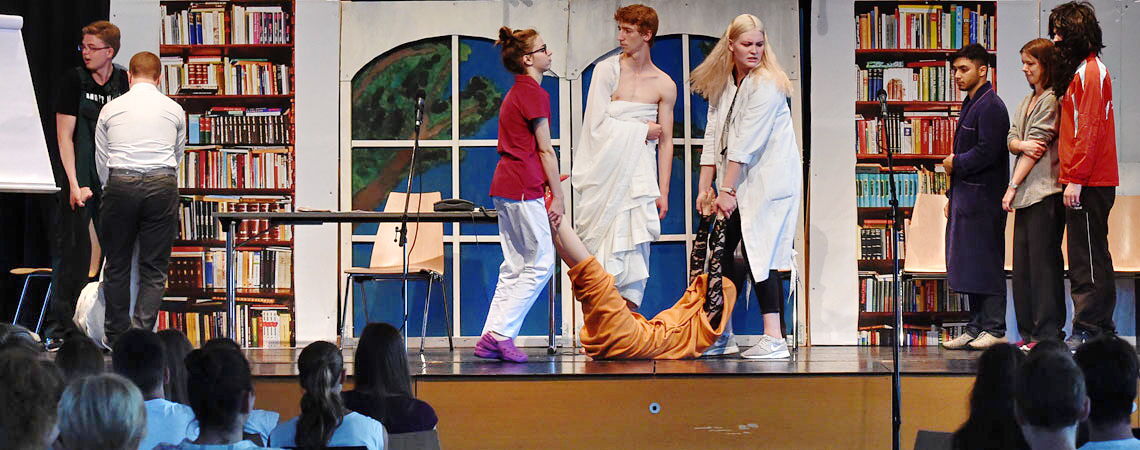 Theateraufführung an der Erich-Bracher-Schule. Mehrere Schülerinnen und Schüler stehen auf der Bühne in verschiedenen Kostümen vor Publikum.