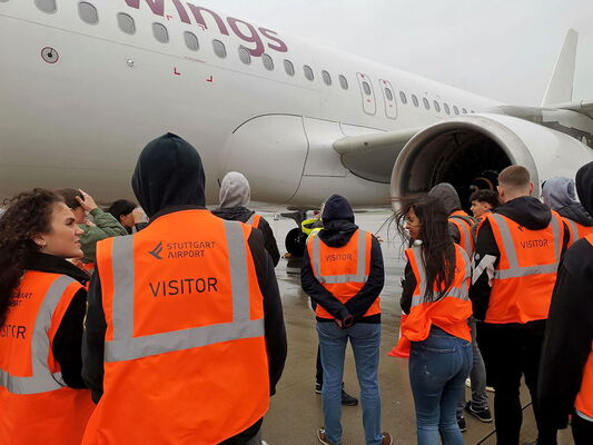Besuchergruppe mit orangefarbenen Warnwesten steht auf dem Rollfeld des Stuttgarter Flughafens vor einem geparkten Flugzeug.