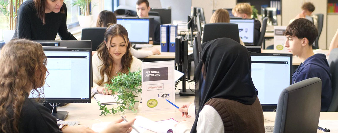 Schülerinnen und Schüler arbeiten in der Übungsfirma des Berufskollegs an Computern. Auf dem Tisch steht ein Schild mit der Aufschrift „Personalabteilung“.